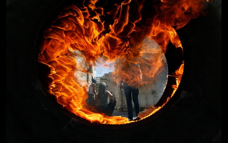 Manifestantes palestinos se ven a través de una llanta incendiada durante enfrentamientos con soldados israelíes en Hebrón, Cisjordania. AFP/H. Bader